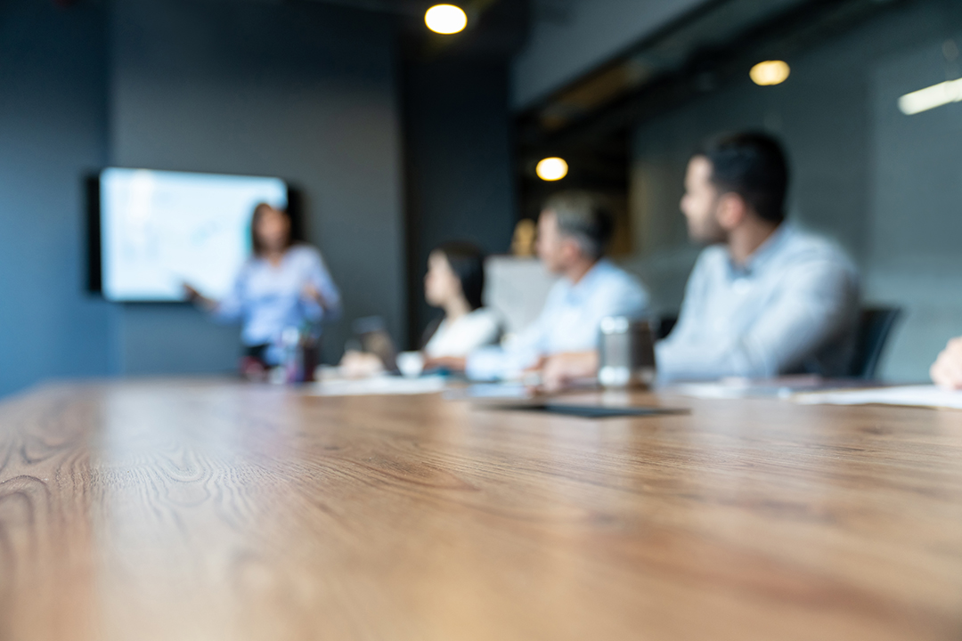 Out of focus image of a woman standing at the head of a conference table presenting something on a TV monitor. There are three people facing her with a fourth's hands just visible.
