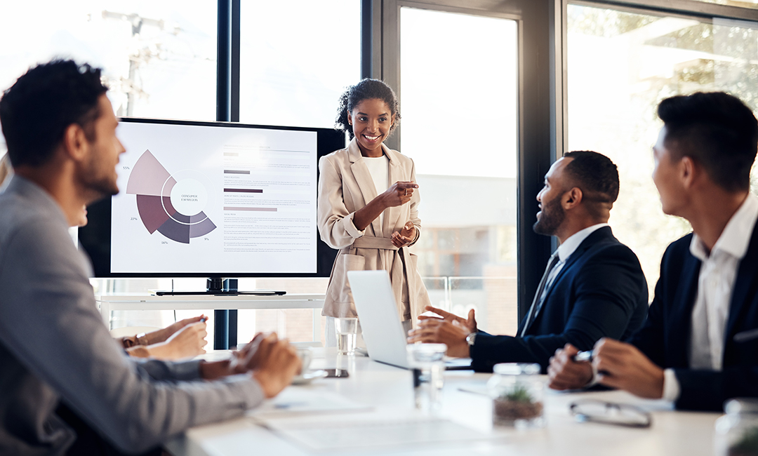 A woman presents data on a TV monitor to a conference table of at least four people.