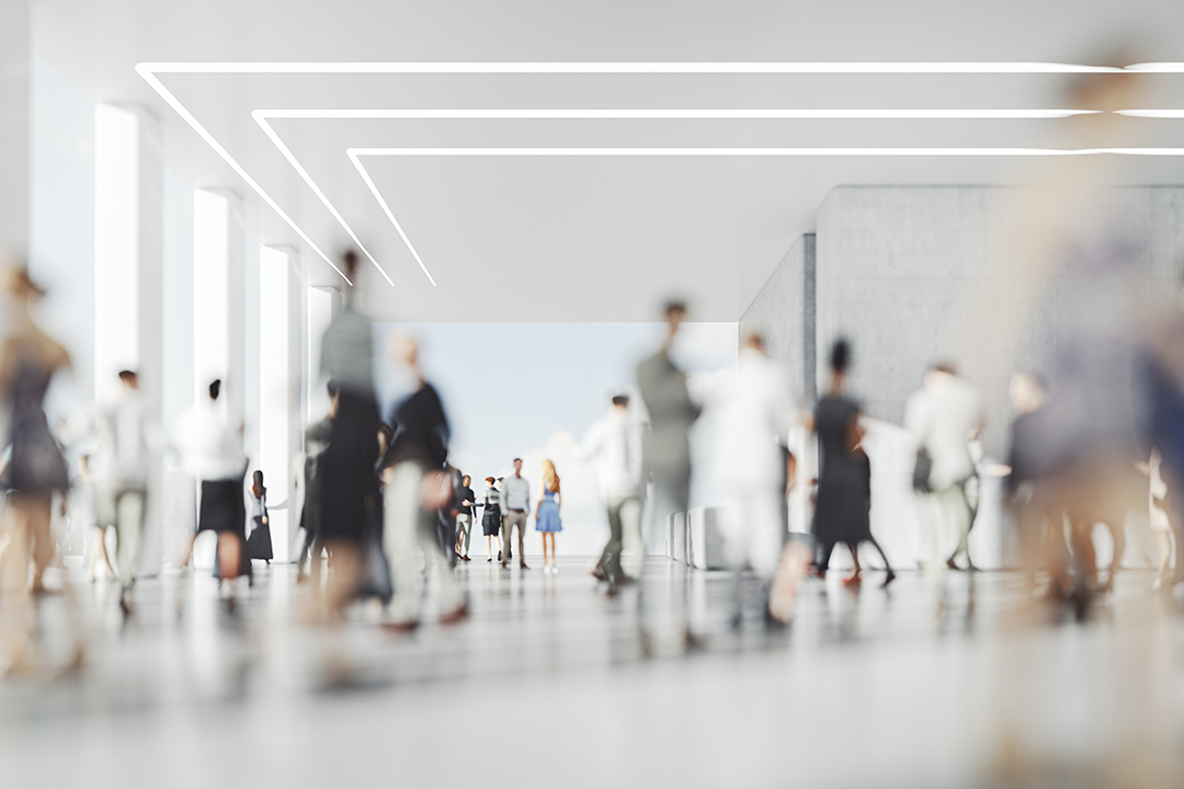 A crowd is out of focus in a large, white room with high ceilings, like an exhibit hall. A cluster of figures at the far side of the room is slightly more in focus.