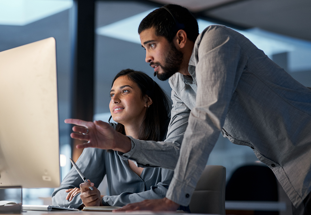 A man points at a computer monitor over a woman's shoulder who is seated in front of him.