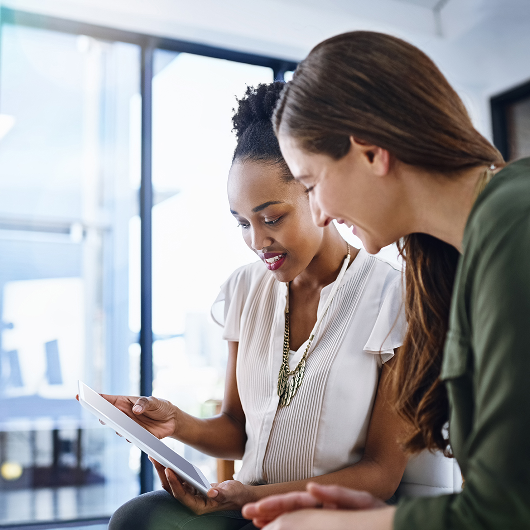 Two women sit together and look at a tablet screen.